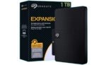 Seagate Expansion portable hard drive beside its black striped packaging, showing 1TB capacity and the Seagate logo on the box and drive surface.