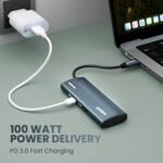 Blue USB-C PD 3.0 charger delivering 100W to a laptop via a USB-C cable, plugged into a wall outlet; text '100 WATT POWER DELIVERY' visible.