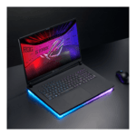 Gaming laptop with RGB-lit base and neon keyboard, open on a dark desk with a wireless mouse nearby (ROG Strix logo on screen).