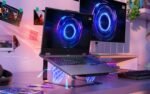 Slim Lenovo laptop on a glass stand with RGB keyboard, connected to two monitors showing neon circular graphics in a vibrant desk setup