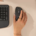 A person’s hand with gold rings using a dark wireless mouse beside a keyboard on a light desk