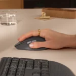 Person's hand with a gold ring resting on a gray wireless computer mouse, with a keyboard in the foreground on a light desk.