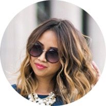 Portrait of a woman with wavy light brown hair, wearing oversized sunglasses and a chunky necklace, looking at the camera.