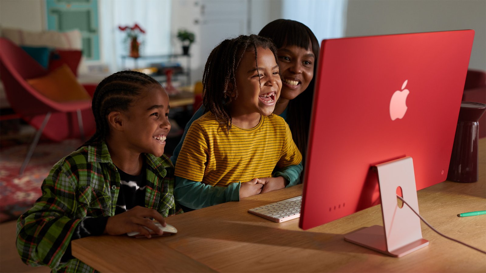 Three kids smile and lean in to view a bright red Apple iMac on a desk, learning together at home. | Ahoundjuè Technologies Three kids smile and lean in to view a bright red Apple iMac on a desk, learning together at home.